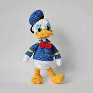 Op de afbeelding: Gehaakte Donald Duck pop. De pop is voornamelijk blauw, wit en geel, met een rode strik en een blauwe matrozenmuts. De pop heeft zwarte ogen en een gele snavel. De pop staat op een witte ondergrond.