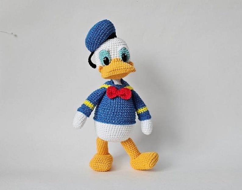 Op de afbeelding: Gehaakte Donald Duck pop. De pop is voornamelijk blauw, wit en geel, met een rode strik en een blauwe matrozenmuts. De pop heeft zwarte ogen en een gele snavel. De pop staat op een witte ondergrond.