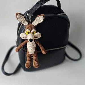 May include: A crocheted Wile E. Coyote keychain in brown, beige, and yellow, hanging in front of a black backpack. The cartoon character has large yellow eyes, a white muzzle, and a black nose. The keychain is attached to a silver-colored chain.