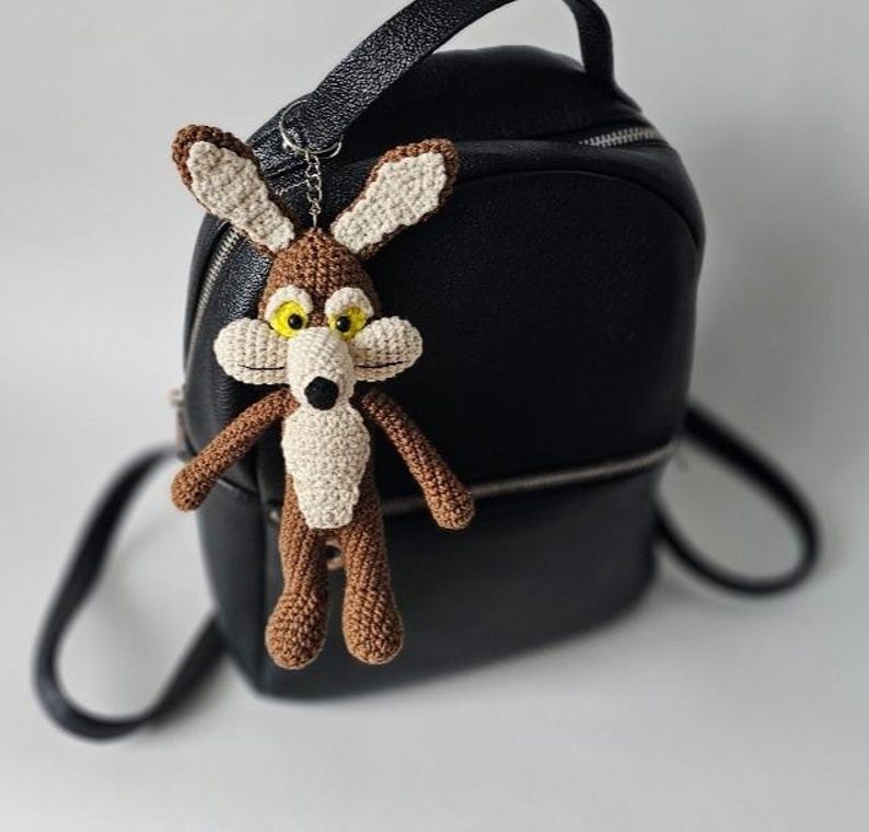 May include: A crocheted Wile E. Coyote keychain in brown, beige, and yellow, hanging in front of a black backpack. The cartoon character has large yellow eyes, a white muzzle, and a black nose. The keychain is attached to a silver-colored chain.
