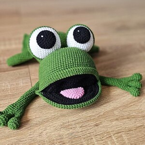 May include: A green crocheted frog toy with large white eyes and a black mouth. The frog has a pink tongue sticking out.
