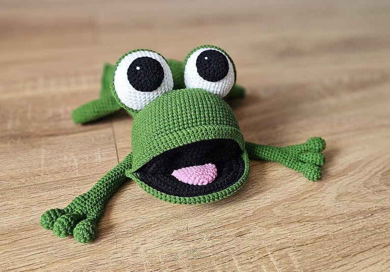 May include: A green crocheted frog toy with large white eyes and a black mouth. The frog has a pink tongue sticking out.