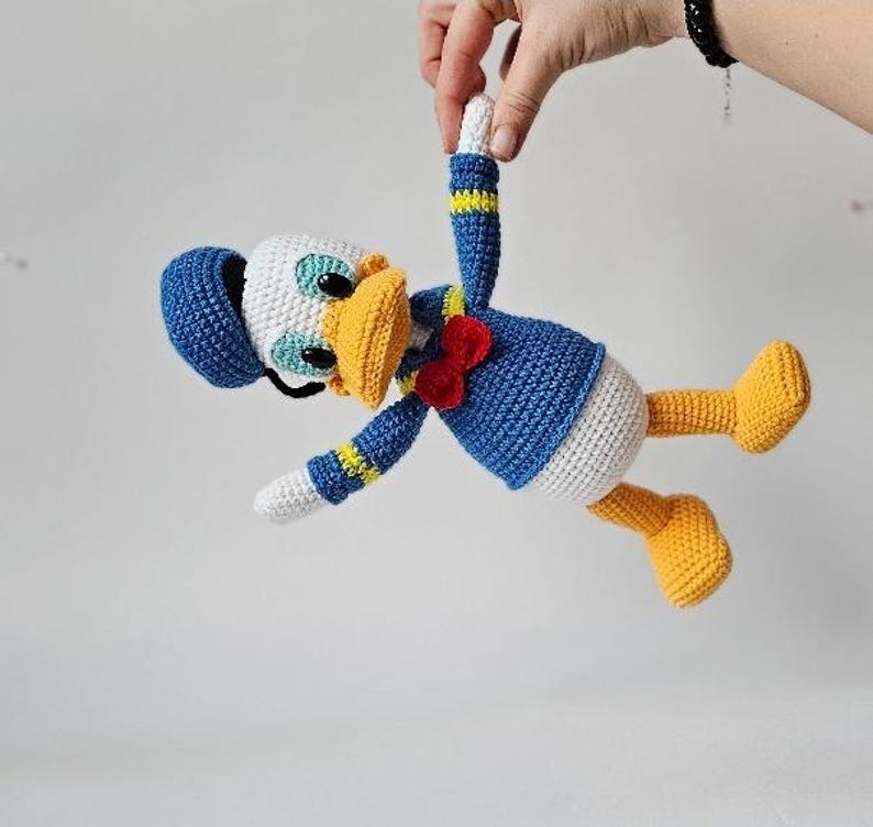 Op de afbeelding: Een handgemaakte gehaakte Donald Duck pop. De pop heeft een blauw matrozenhemd, een witte buik, gele voeten en een gele snavel. De pop heeft een blauwe hoed en een rode vlinderdas. De pop wordt door een hand omhoog gehouden.