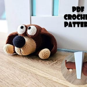 Puede incluir: Un tope de puerta con forma de perro tejido a ganchillo en marrón, beige y negro. El perro tiene ojos blancos grandes, una nariz negra y patas beige. El texto "PDF CROCHET PATTERN" se muestra encima del perro. Se muestra una imagen más pequeña del tope.