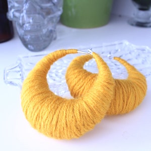 May include: A pair of large, yellow yarn hoop earrings. The earrings are made of a thick, yellow yarn that is wrapped around a metal hoop.