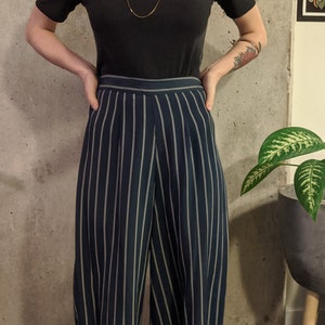 May include: A black ribbed short-sleeve top and navy blue and white striped wide-leg pants. A long, delicate gold necklace is worn. The outfit is styled against a concrete wall.