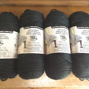 May include: Four skeins of black Soft & Shiny yarn by Loops & Threads. Each label displays the text Soft & Shiny, Doux et Brillant, Suave y Brillante, and indicates a weight of 6 oz and 170 g, with a length of 311 yards and 285 meters per skein.