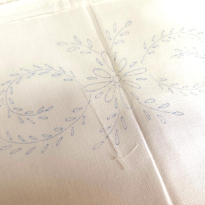 Peut inclure: Un morceau de tissu blanc cassé présentant un motif de transfert bleu clair composé d'une fleur centrale entourée de deux motifs de vignes symétriques. Le dessin est formé de petites lignes en pointillés pour la broderie.