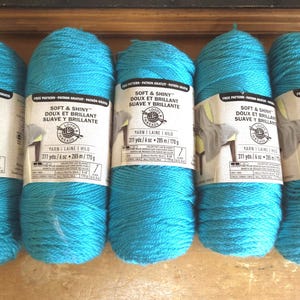 Può includere: Cinque gomitoli di filato turchese brillante, ciascuno avvolto in un'etichetta di carta con la scritta Soft & Shiny, Loops & Threads, 311 yds, 6 oz, 285 m e 170 g. Il filato ha una consistenza liscia e leggermente riflettente.