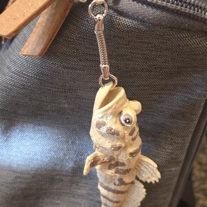 May include: A keychain featuring a small, cream-colored fish figurine with brown horizontal stripes and a wide-open mouth. The fish is attached to a silver-toned metal split ring by a short, flexible chain, showcasing detailed scales and a single visible eye.