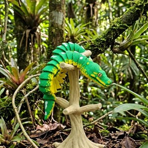 May include: A green and yellow articulated crocodile figurine with a segmented body, resting on a beige, textured tree-shaped stand. The crocodile features a detailed scale pattern, a pointed snout, and visible eyes, showcasing a flexible, jointed design.