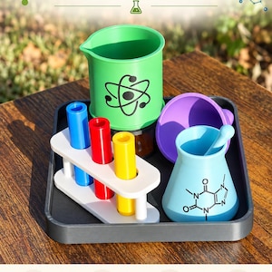 Peut inclure: Un ensemble de jouets sur le thème scientifique imprimés en 3D sur un plateau sombre, comprenant un bécher vert avec une icône d'atome, un support à tubes à essai blanc avec des tubes bleu, rouge et jaune, un entonnoir violet et un flacon bleu clair.