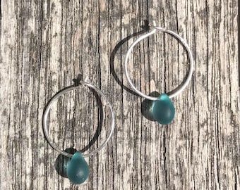 May include: Two silver hoop earrings, each with a teal teardrop-shaped glass bead. The earrings are set against a weathered wooden backdrop, highlighting their simple design and natural appeal. The beads are a lovely shade of teal.