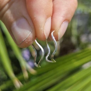 May include: A pair of silver wave-shaped earrings. The earrings have a hammered texture and a curved design, resembling a gentle wave. The earrings are held against a blurred green background.