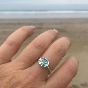 May include: A silver ring with an oval abalone shell inlay, displaying iridescent blues, greens, and purples. The ring is set against a blurred beach backdrop, emphasising the natural colours of the ocean.