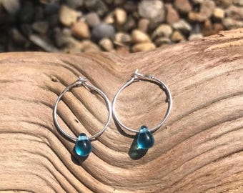 May include: Silver hoop earrings, each featuring a teal glass teardrop bead. The earrings are presented on a piece of weathered wood, with a blurred background of small stones.