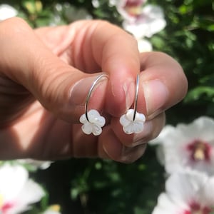 Op de afbeelding: Een paar zilveren creolen met witte, bloemvormige bedels. De oorbellen zijn gemaakt van metaal en hebben een eenvoudig ontwerp.