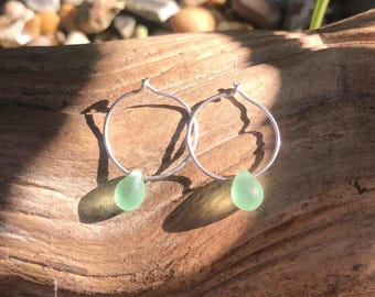 May include: A pair of silver hoop earrings with a light green sea glass teardrop charm hanging from each hoop.