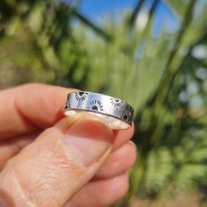 May include: A silver ring with a repeating sunburst pattern. The ring is held between fingers, with a blurred green and blue background. The ring's design features small suns with rays, creating a continuous band around the ring.