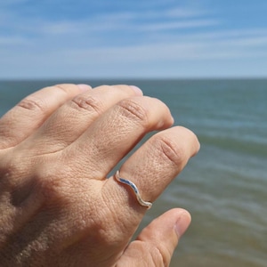 Może przedstawiać: Srebrny pierścionek o falistym wzorze. Pierścionek jest noszony na palcu, z błękitnym niebem i morzem w tle. Design pierścionka przypomina delikatną falę.