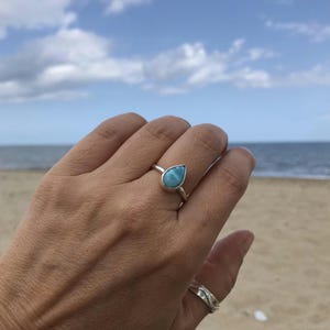 May include: A silver ring with a pear-shaped, light blue gemstone. The ring is set against a beach background with a blue sky and ocean. The ring is on a hand.