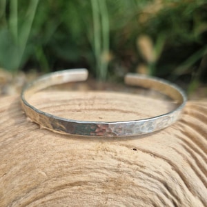 Könnte beinhalten: Ein silbernes Armreif-Armband mit gehämmertem Finish. Das Armband hat eine strukturierte Oberfläche mit einem gehämmerten Design. Das offene Manschettendesign ermöglicht ein einfaches Tragen. Das Armband wird auf einer Holzoberfläche gezeigt.