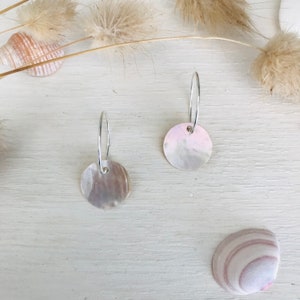 May include: A pair of silver hoop earrings with a round, iridescent shell pendant. The earrings are on a white surface with a seashell in the background.
