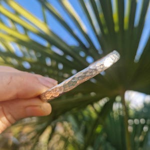 May include: A silver-coloured, hammered metal bracelet. The bracelet has a textured surface with small, irregular indentations. The bracelet is held against a background of green leaves and blue sky.