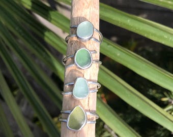May include: Four silver rings are arranged vertically on a wooden stick. Each ring features a smooth, rounded piece of sea glass in varying shades of green, ranging from pale mint to a deeper olive tone, set within a simple metal band.
