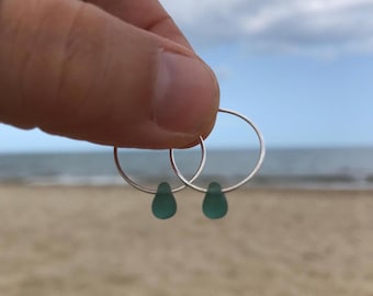 May include: Silver hoop earrings with teal teardrop pendants. The earrings are displayed against a beach scene with a sandy shore, sea, and a cloudy sky in the background.