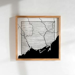 Pode incluir: Uma ilustração em preto e branco emoldurada de uma costa com um fundo texturizado. A ilustração é emoldurada em uma moldura de madeira natural.