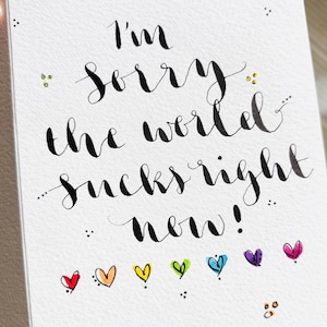 K&ouml;nnte beinhalten: Eine wei&szlig;e Karte mit schwarzer Kalligrafie, auf der "Es tut mir leid, die Welt ist im Moment schei&szlig;e!" steht, mit einer Reihe bunter Herzen am unteren Rand.