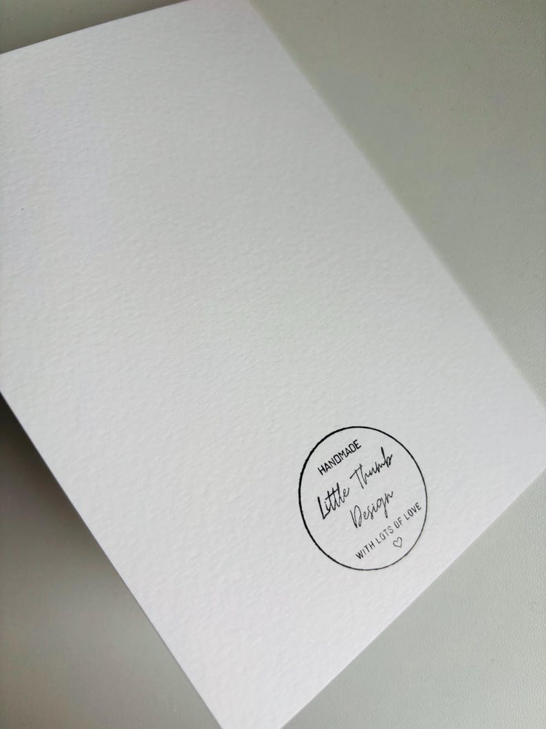 K&ouml;nnte beinhalten: Eine wei&szlig;e Karte mit einem schwarzen runden Stempel, auf dem "Handmade Little Thumb Design with lots of love" in handschriftlicher Schrift steht.