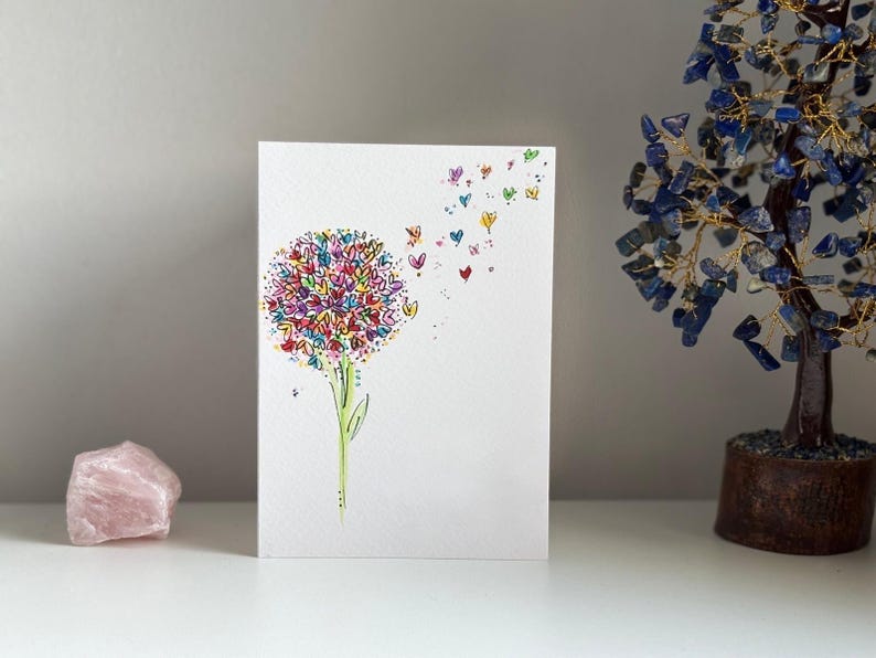 K&ouml;nnte beinhalten: Eine wei&szlig;e Karte mit einer bunten Illustration einer Blume mit Herzen als Bl&uuml;tenbl&auml;tter. Die Herzen fliegen auch von der Blume weg.