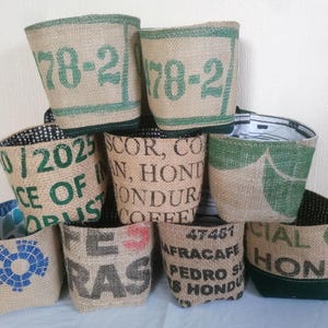 May include: A selection of burlap planters, each with unique printed designs and text. The planters have green and black trim, with some displaying numbers and phrases such as "78-2" and "CE OF OPUS". The interiors are lined with patterned fabric.
