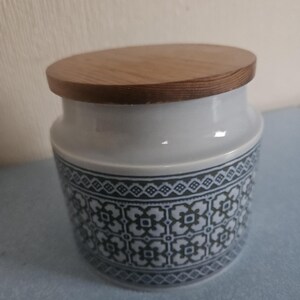 May include: A white ceramic jar with a wooden lid. The jar has a dark blue geometric pattern around the base. The lid is round and made of light-coloured wood. The jar is designed for storage.