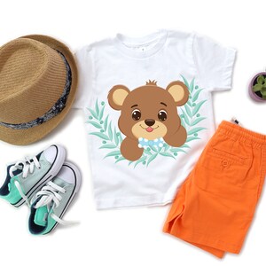 K&ouml;nnte beinhalten: Ein wei&szlig;es T-Shirt mit einem braunen B&auml;ren, der eine blaue Schleife tr&auml;gt, in einem gr&uuml;nen Bl&auml;tterrahmen. Der B&auml;r schaut den Betrachter an. Das Shirt wird mit orangefarbenen Shorts und gr&uuml;n-blauen Turnschuhen kombiniert.