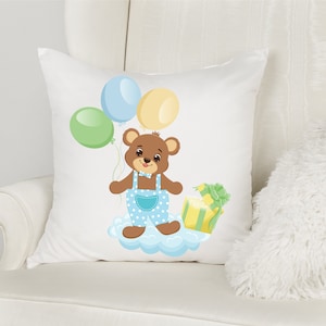 K&ouml;nnte beinhalten: Ein wei&szlig;es Kissen mit einem braunen B&auml;ren, der eine blaue Latzhose und eine Fliege tr&auml;gt. Der B&auml;r h&auml;lt ein gr&uuml;n-gelbes Geschenk und ist von blauen, gr&uuml;nen und gelben Luftballons umgeben.