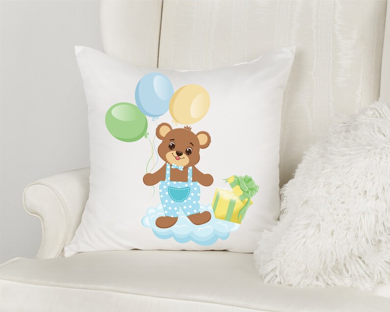 K&ouml;nnte beinhalten: Ein wei&szlig;es Kissen mit einem braunen B&auml;ren, der eine blaue Latzhose und eine Fliege tr&auml;gt. Der B&auml;r h&auml;lt ein gr&uuml;n-gelbes Geschenk und ist von blauen, gr&uuml;nen und gelben Luftballons umgeben.