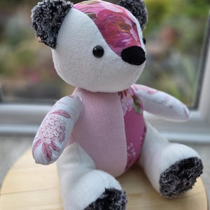 May include: A stuffed bear toy constructed from various fabric panels. The bear features white, pink, and dark grey fuzzy textures, with a floral pattern on the head and torso. It has black button eyes and a black nose, with dark grey fuzzy ears and feet.