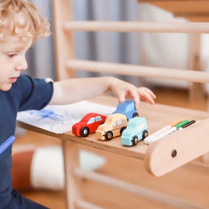 May include: A young child sits at a wooden table with a white sheet of paper and colorful markers.  The child is drawing on the paper while looking at the markers.  There are four small wooden toy cars on the table in front of the child.