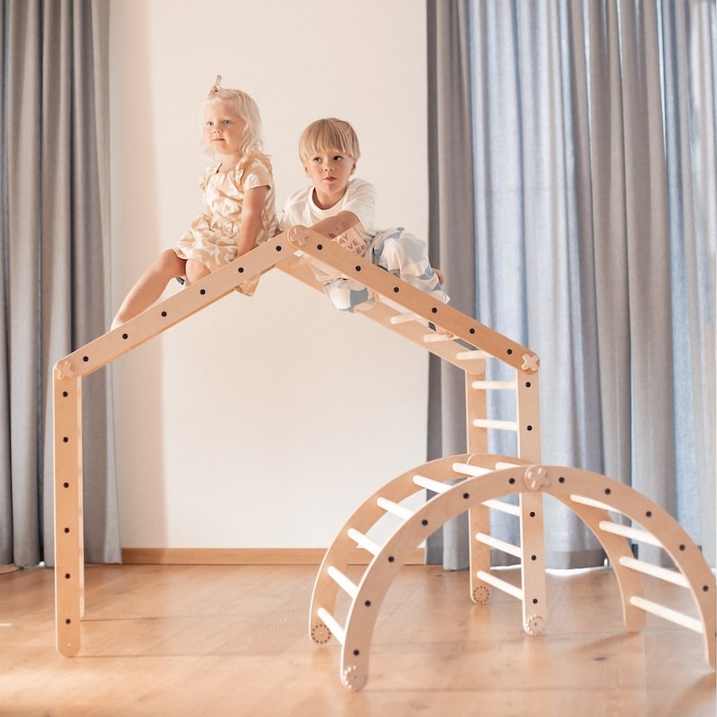 May include: Two children are playing on a wooden climbing structure. The structure includes a climbing wall, a ladder, and a curved archway. The children are smiling and having fun.