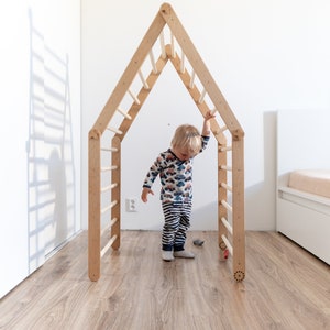 May include: A young child stands inside a wooden play structure shaped like a house. The structure has a ladder on each side and a small opening at the top. The child is wearing a colorful shirt and striped pants.