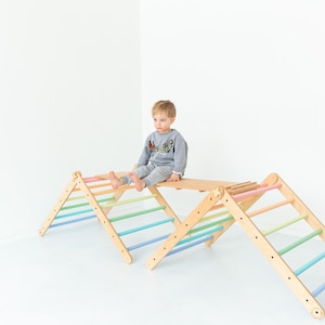 May include: A wooden climbing triangle with rainbow colored rungs. A child is sitting on the top platform of the triangle.