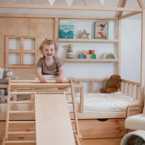 May include: A child sits on a wooden climbing frame with a slide attached. The frame is part of a wooden playhouse with shelves and a bed. The playhouse is decorated with a string of flags and a book titled "Happiness".