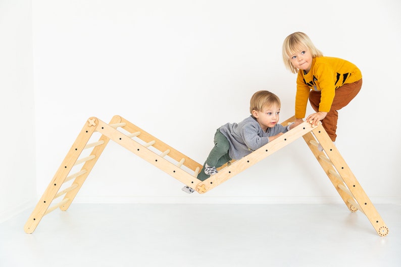 May include: Two children play on a wooden climbing frame with a ladder. The frame is made of natural wood and has a triangular shape. The children are both wearing casual clothing.