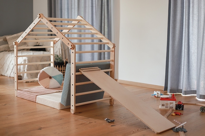 May include: A wooden play structure with a climbing ladder, a slide, and a padded floor. The structure is painted in natural wood tones and has a small house-like design.
