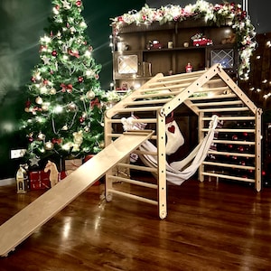 Op de afbeelding: Houten speelstructuur met een glijbaan, een klimmuur en een hangmat. De structuur is gemaakt van natuurlijk hout en heeft een lichtbruine afwerking. De glijbaan is bevestigd aan de zijkant van de structuur en is gemaakt van glad, licht hout. De klimmuur is bevestigd aan de voorkant van de structuur en is gemaakt van stevige, natuurlijke houten spijlen. De hangmat is bevestigd aan de bovenkant van de structuur en is gemaakt van zachte, witte stof. De structuur is opgezet in een kamer met een kerstboom op de achtergrond.