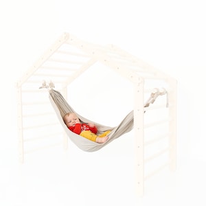 May include: A white wooden climbing frame with a gray fabric hammock hanging from it. A baby is lying in the hammock.