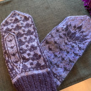 May include: A pair of purple and white knitted mittens with a geometric pattern. The mittens are folded in half and are lying on a green fabric surface.